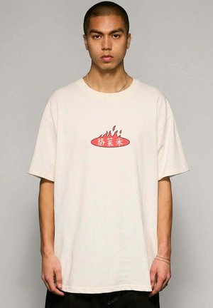 Młody mężczyzna z krótkimi włosami, ubrany w luźny biały T-shirt z czerwonym płonącym owalem i azjatyckimi znakami na przodzie, stoi na szarym tle.