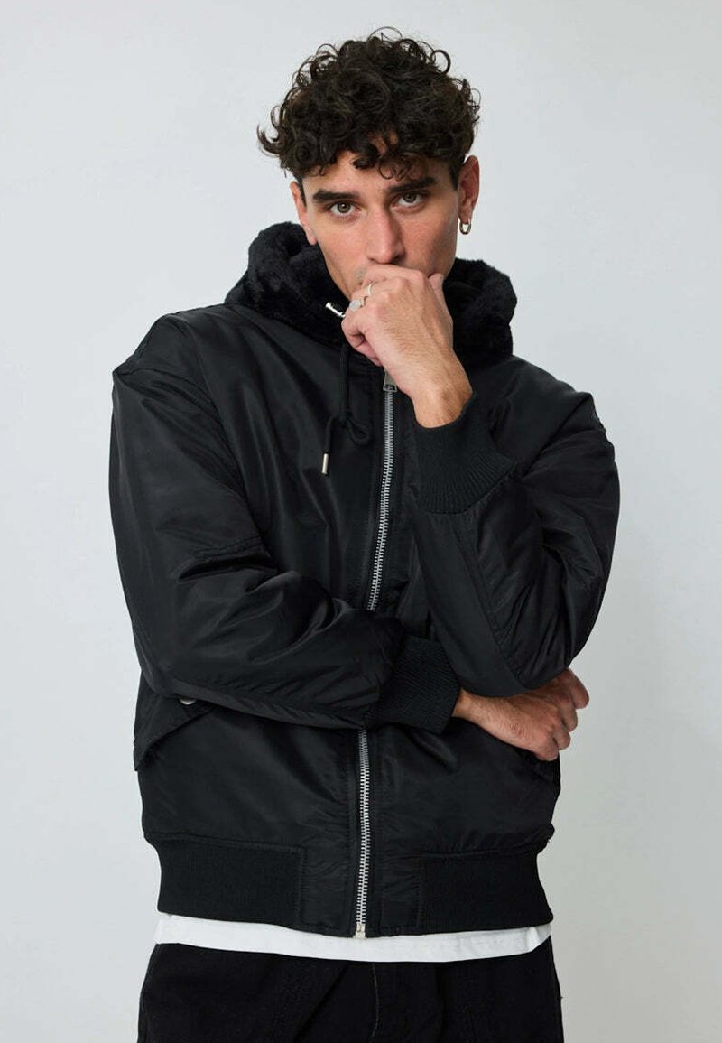 Veste bomber noire avec une capuche doublée en fourrure, fermeture éclair sur le devant, poignets côtelés et ourlet. Présente une texture lisse et une coupe décontractée.