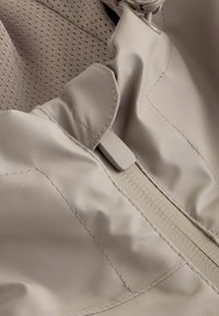 Gros plan sur un tissu de veste beige avec une fermeture éclair et une doublure en mesh visibles dans le coin supérieur gauche.