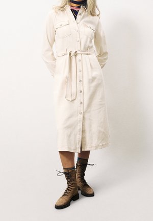 Dlouhé béžové košilové šaty s límečkem, zapínáním na knoflíky vpředu a dvěma náprsními kapsami. Nošené s hnědými šněrovacími botami a látkovým páskem uvázaným na uzel. Hladká textura.