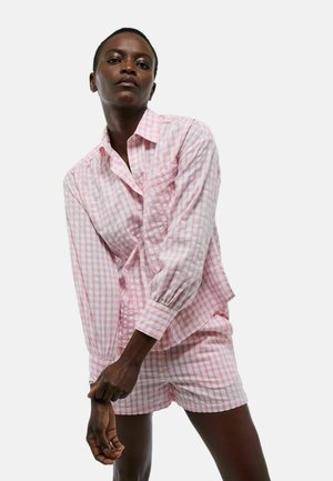 Personne portant une chemise à manches longues à carreaux roses et blancs et un short assorti, sur fond blanc uni.