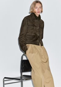 Bruine suède jas met kraag, knopen aan de voorkant en twee zakken, gecombineerd met beige broek met wijde pijpen. Minimalistisch ontwerp tegen een effen achtergrond.