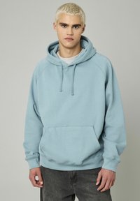 Jeune homme aux cheveux blonds courts portant un sweat à capuche bleu clair et un jean foncé, se tenant devant un fond gris uni.