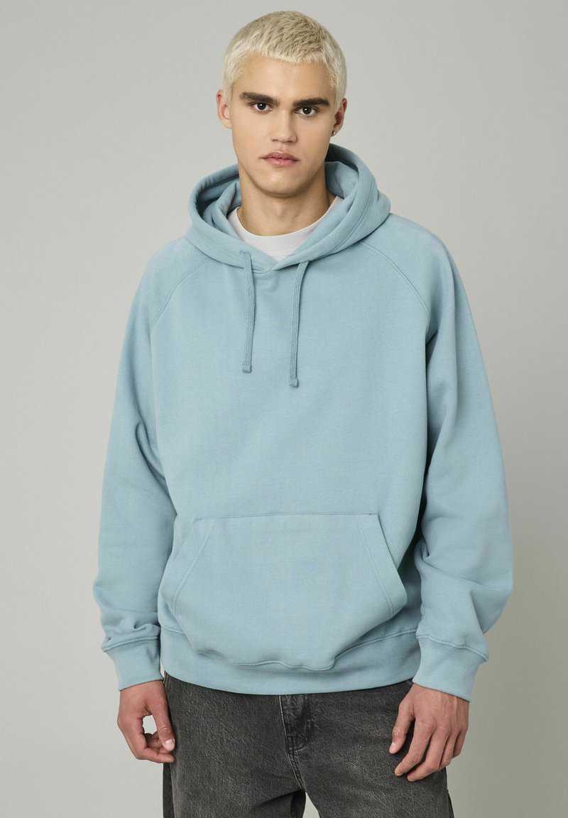 Jeune homme aux cheveux blonds courts portant un sweat à capuche bleu clair et un jean foncé, se tenant devant un fond gris uni.