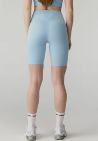 Femme portant un short cycliste taille haute bleu clair, des chaussettes blanches avec texte noir, et des baskets blanches, debout de dos sur un fond uni.