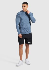 Hellblaues langärmliges Sportshirt mit Raglanärmeln, kombiniert mit schwarzen Shorts mit einem weißen Logo. Weiße Sneakers runden das Outfit ab.