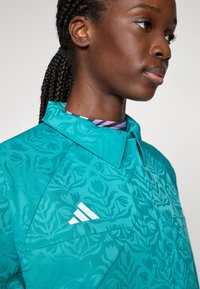 Kvinna med flätat hår som bär en turkosmönstrad jacka med Adidas-logotyp, visad i en närbild av sidan.