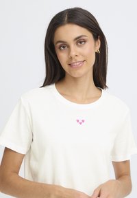Fehér, rövid ujjú pamut póló, a mellkasán három rózsaszín hímzett szívvel. Sima textúra és klasszikus kerek nyakkivágás.