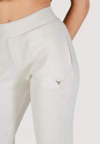 Pantaloni bianchi a sigaretta con una texture liscia, dotati di una tasca laterale e di un accento in metallo triangolare sul coscia sinistra.
