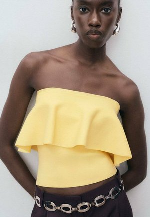 Jonge vrouw in een gele strapless gerimpelde top, donkere broek met een zilveren schakelriem en grote oorbellen, staande tegen een lichte muur.