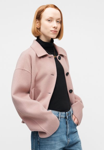 Νεαρή γυναίκα με ανοιχτό κόκκινο καρέ κούρεμα, φορώντας ροζ παλ oversized σακάκι με κουμπιά, μαύρο ζιβάγκο, μπλε τζιν και χρυσό κολ ιέ.