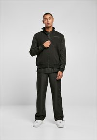 Svart zip-up jakke med høy krage, kombinert med matchende bukser. Begge har en jevn tekstur og minimalistisk design. Brukt med hvite joggesko.