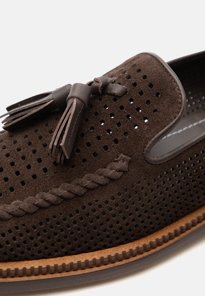 Mocassin en daim marron avec design perforé, bordure tressée et pampilles en cuir sur une semelle marron clair.