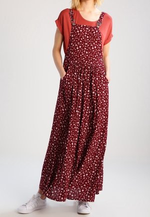 Kobieta nosząca rdzawo-pomarańczową koszulkę z krótkim rękawem pod długą bordową sukienkę na szelkach w kwiatowy wzór z kieszeniami, zestawioną z białymi trampkami.