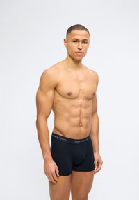 Άνδρας με κοντά μαλλιά, όρθιος, φοράει μαύρα εσώρουχα Tommy Hilfiger boxer, μπροστά σε απλό ανοιχτό γκρι φόντο.