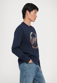 Sweatshirt azul-marinho com decote redondo, punhos em ribana, apresentando um logótipo estilizado em tons de laranja e bege na parte da frente. Corte casual.