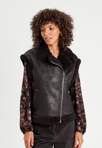 Gilet in finta pelle nera con colletto e orlo in finta pelliccia. Maniche lunghe a fantasia in tonalità scure con accenti floreali. Chiusura frontale con zip.