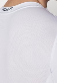 Vit sporttröja i strukturerat, ventilerande material, med rund halsringning och en liten logotyp vid kragen.