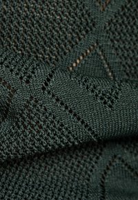 Tela tejida de color verde oscuro con un patrón geométrico de diamantes; presenta una superficie texturizada con secciones de punto abierto y otras lisas.