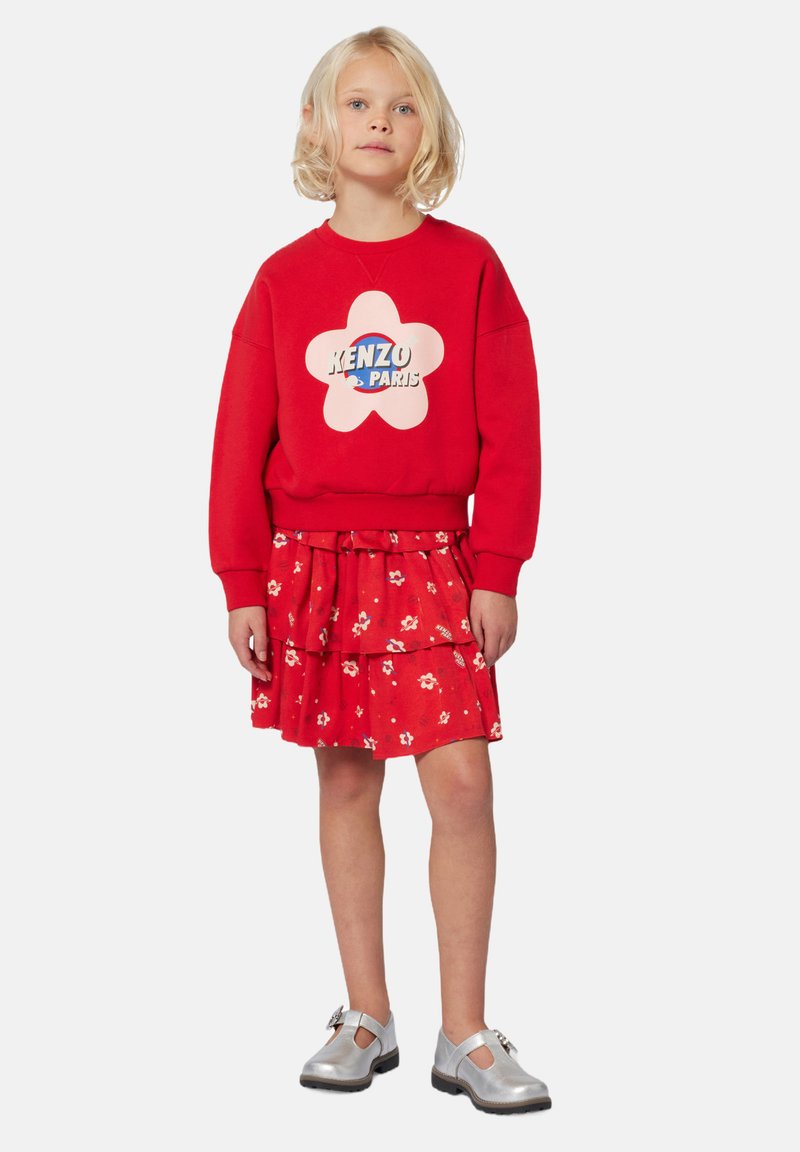 Rode sweatshirt met een roze bloemenlogo en de tekst "KENZO PARIS," gecombineerd met een rode gelaagde rok met kleine witte bloemenpatronen. Zilveren schoenen.