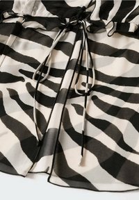 Blouse noire et beige avec un imprimé zèbre ondulé, dotée d'un cordon de serrage à la taille et d'une texture de tissu léger et transparent.