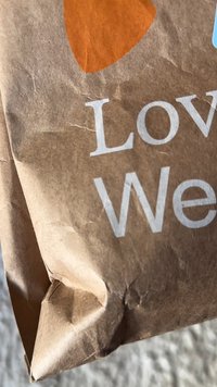 Braune Papiertüte mit zerknitterter Textur, versehen mit einem orangefarbenen Herzgrafik und weißem Text "Liebe" und "Wir". Die obere Ecke ist leicht eingerissen.