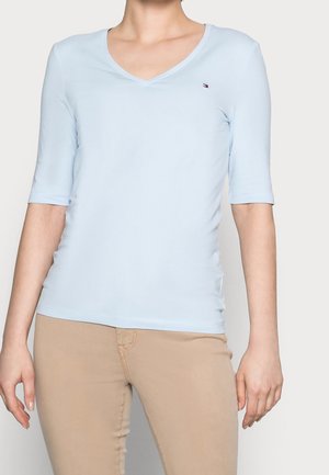Femme portant un haut bleu clair à col en V et manches courtes et un pantalon beige, debout devant un fond clair uni.