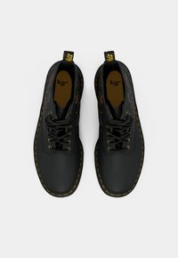 Un paio di stivali neri Dr. Martens con cuciture gialle e ricami floreali colorati sui lati, visti dall'alto su sfondo bianco.