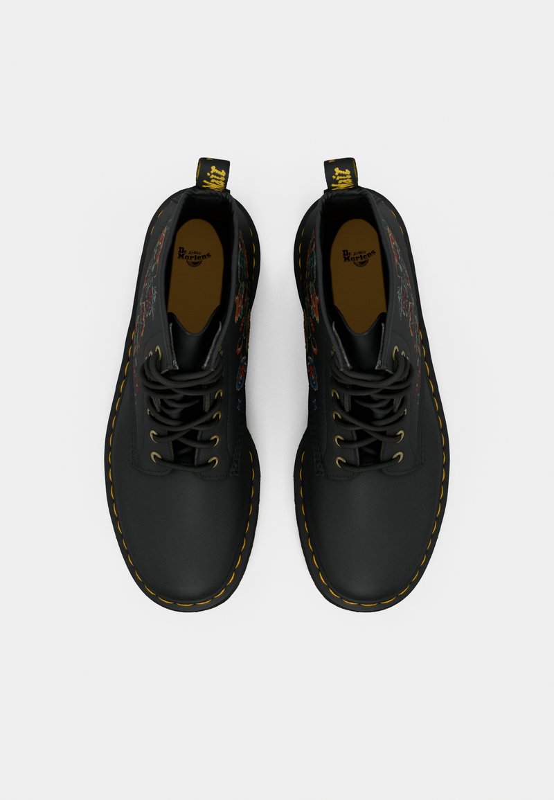 Une paire de bottes Dr. Martens noires avec des coutures jaunes et des broderies florales colorées sur les côtés, vue de dessus sur un fond blanc.
