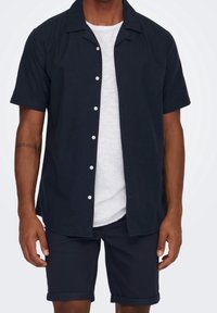 Camicia button-up a maniche corte scura con colletto da campeggio, adornata da bottoni bianchi, abbinata a pantaloni corti scuri arrotolati e una maglietta bianca sottostante.