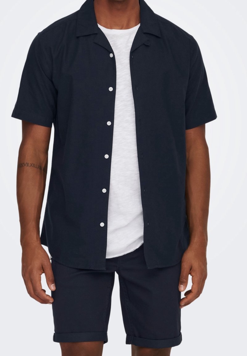 Camicia button-up a maniche corte scura con colletto da campeggio, adornata da bottoni bianchi, abbinata a pantaloni corti scuri arrotolati e una maglietta bianca sottostante.