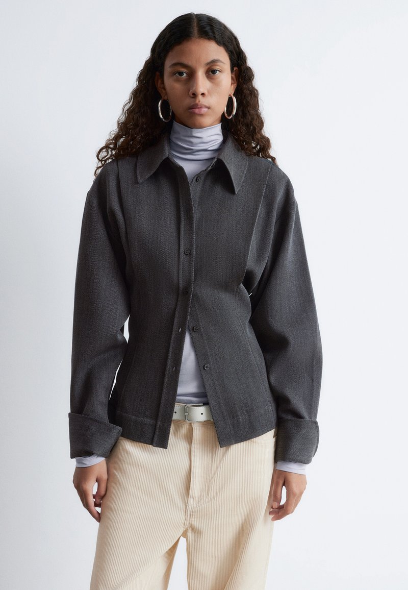 Gray button-up shirt with a textured fabric, pointed collar, and long sleeves, paired with a light purple turtleneck and cream-colored wide pants.