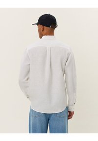 Camicia bianca in lino con maniche lunghe, dettaglio delle pieghe sul retro e orlo arrotondato. Indossata con pantaloni in denim blu e berretto blu navy.