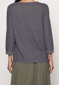 Grauer gerippter Pullover mit langen Ärmeln und umgeschlagenen Bündchen. Weicher Stoff, lockere Passform und runder Halsausschnitt. Über einem grünen Rock getragen.