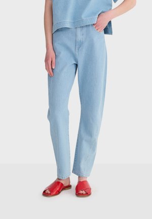 Osoba ubrana w jasnoniebieskie jeansy z wysokim stanem, dopasowany krótki top w tym samym odcieniu niebieskiego oraz czerwone sandały na płaskiej podeszwie z odkrytymi palcami, stojąca z jedną ręką w kieszeni.