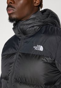 Jachetă puffer neagră cu guler înalt și închidere cu fermoar. Are o textură distinctă și un logo alb pe pieptul stâng.