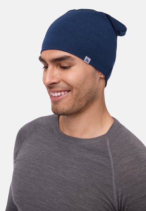 Navy blue knitted beanie with a pointed top and a small logo tag. Worn by a person in a grey long-sleeve shirt.