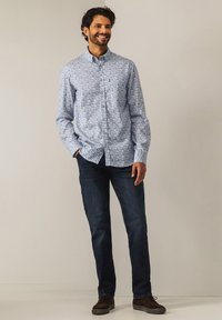 Hemd mit Knopfleiste aus hellblauem Baumwollstoff mit floralem Muster, kombiniert mit dunkelblauen Jeans und braunen Wildlederschuhen, vor einem neutralen Hintergrund stehend.