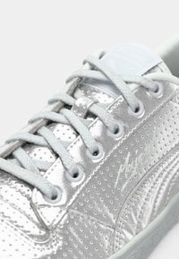 Sølvmetallic sneakers med hvide snørebånd, perforerede sider og præget "Puma" og "Maje" logoer på en hvid baggrund.