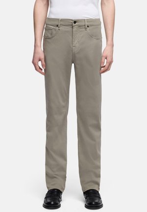 Man die beige rechte broek, wit overhemd ingestopt en zwarte nette schoenen draagt, staand tegen een effen witte achtergrond.