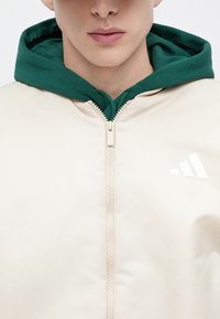 Tõmblukuga heleroosa jope, mille all roheline kapuuts, rinnal valge kolmnurkne logo ning siledast, läikivast materjalist.