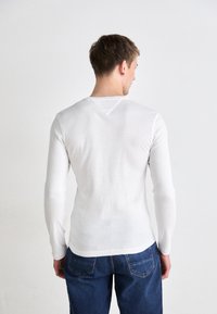 Vit långärmad skjorta i lätt tyg, med rund halsringning och en triangulär sömdetalj på baksidan, kombinerad med blå jeans.