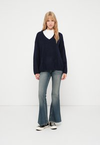 Szőke nő áll, sötétkék V-nyakú pulóvert, fehér alját, halvány farmernadrágot és fekete alacsony top cipőt visel, fehér háttér előtt.