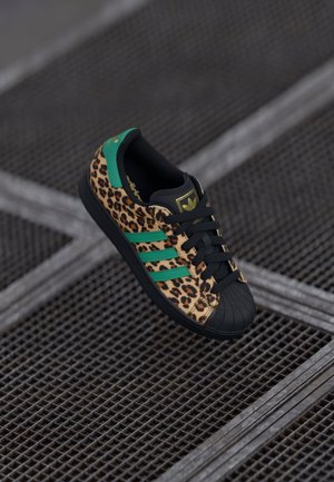 Adidas sneaker met luipaardprint, groene strepen, zwarte teenkap en veters, liggend op een metalen rasteroppervlak.
