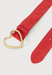 Cinto em camurça vermelha com fivela dourada em forma de coração. O cinto tem uma textura suave e uma extremidade afunilada com um único furo de ajuste.