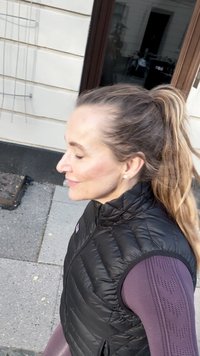 Czarna puchowa kamizelka z wysokim kołnierzem na długim rękawie fioletowej bluzki z wzorzystymi rękawami. Jasnobrązowe włosy w kucyku.