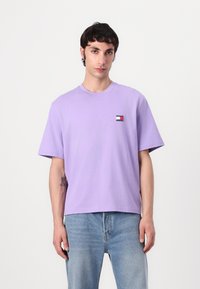 Purple cotton t-shirt with short sleeves and a crew neck. Includes a small embroidered logo patch featuring red, white, and blue colors on the chest.