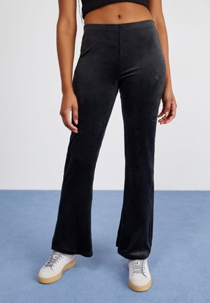 Woman wearing black velour flare pants, white sneakers with beige soles, and a cropped black top, standing on blue floor against white wall.