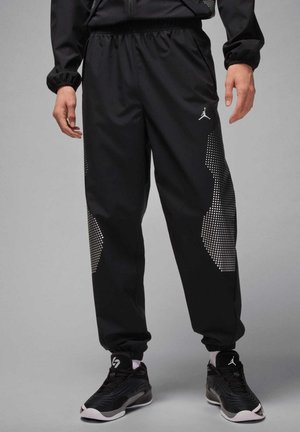 Mann trägt schwarze Jogginghose mit weißen gepunkteten Seitenmustern und schwarze Sneaker mit weißen Sohlen und Logos.