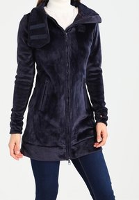 Donna che indossa una lunga giacca in fleece blu navy con zip, dotata di un colletto alto e tasche laterali con zip, abbinata a jeans blu scuro.